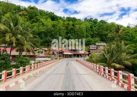 Eine Betonbrücke mit rot-weißem Geländer überquert ein kleines ländliches Dorf, das von dichtem grünen Wald und Palmen im Norden Laos umgeben ist. Traditionelle Holzhäuser mit Metalldächern liegen am Fuße eines üppigen Hügels unter einem hellblauen Himmel mit verstreuten Wolken. Stockfoto