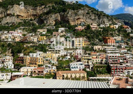 Reihen von bunten Gebäuden in Pastelltönen stürzen sich auf den steilen Hügel von Positano, Italien, vor zerklüfteten Kalksteinklippen und üppigem Grün unter hellem Himmel. Die lebendige Architektur und die mehrschichtigen Terrassen schaffen eine beeindruckende mediterrane Dorffläche. Stockfoto