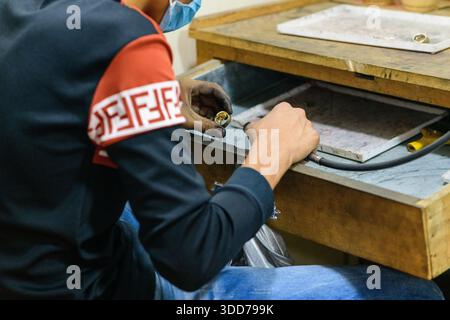 Eine Nahaufnahme der Hände, die an einem Goldring mit einem Drehwerkzeug auf einer Holzwerkbank in Battambang, Kambodscha, arbeiten. Das Bild zeigt den handwerklichen Prozess, die strukturierten Oberflächen und die praktische Umgebung einer traditionellen Schmuckwerkstatt. Stockfoto