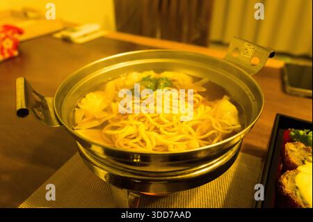 Ein Metalltopf mit dampfenden Nudeln, Knödeln und Gemüse steht auf einem gewebten Tischset, das in warmes goldenes Licht getaucht ist, für eine gemütliche Atmosphäre. Stockfoto