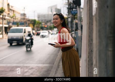 Asiatische Frau lacht, während sie an der Stadtstraße wartet und ihr Smartphone hält Stockfoto