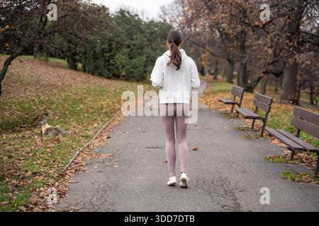 Frau, die im Herbstpark joggt, mit weißem Hoodie. Auf dem Weg durch den friedlichen Stadtpark laufen, aktiver Lebensstil und Selbstpflege im Freien zeigen. Stockfoto