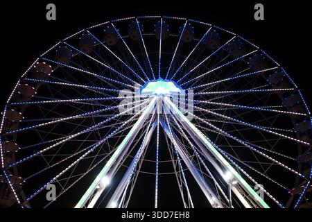 Niedrigwinkelaufnahme eines großen beleuchteten Riesenrads vor einem tiefschwarzen Himmel. Die geometrische Symmetrie der Speichen und das leuchtende Blau und Stockfoto
