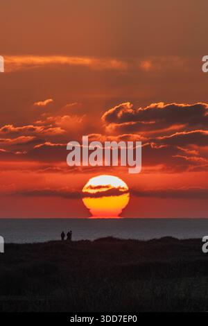 Die Sonne schmilzt im Meer in einem orangefarbenen Leuchten, das hinter fernen Wolken schmilzt .3 Menschen beobachten den Sonnenuntergang von der Landzunge auf dem Küstenpfad in der Nähe von Mumbles, S. Wales. Stockfoto
