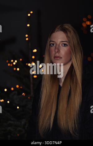 Das ist ein Porträt einer jungen Frau mit langen, fließenden blonden Haaren. Sie hat helle Haut und trägt ein schwarzes Oberteil und eine dunkle Jacke. Ihr Ausdruck ist Stockfoto