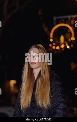 Dieses Foto zeigt eine junge Frau mit langen, fließenden blonden Haaren, die vor einem Hintergrund aus verschwommenen Weihnachtslichtern positioniert ist. Die Beleuchtung ist Stockfoto