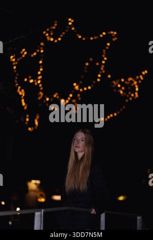 Das Foto zeigt eine junge Frau, die vor einem großen, beleuchteten Weihnachtsstern aus Lichterketten steht. Die Frau ist leicht positioniert Stockfoto