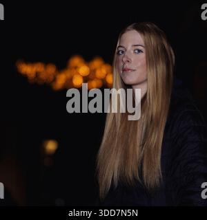 Dieses Foto fängt eine junge Frau während eines Weihnachtsfotos ein. Sie hat lange blonde Haare und trägt eine dunkle Pufferjacke. Der Hintergrund ist Stockfoto
