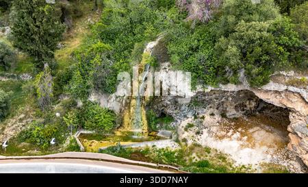 Blick aus der Vogelperspektive auf einen kleinen natürlichen Wasserfall, der über Kalkstein fließt, neben einer Landstraße in Elaiona, Zentralgriechenland, umgeben von grüner Vegetation und Stockfoto