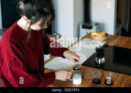 Die junge Frau benutzt Milchaufschäumer an der Holzküche neben Kaffeetassen. Kaffeespezialitäten, Barista-Fertigkeiten, Bohnen mit einmaliger Herkunft, Kunsthandwerk Stockfoto