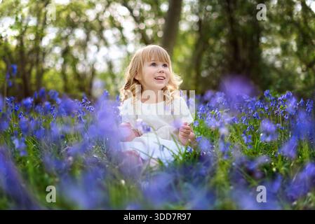 Glückliches Mädchen, das zwischen blühenden Glockenblöcken auf der Waldlichtung sitzt. Stockfoto
