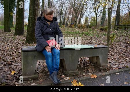 Eine ältere Frau, die allein auf einer Betonparkbank sitzt und ein rotes Buch hält, umgeben von Herbstlaub Stockfoto