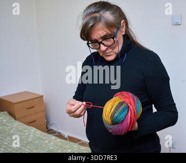 Seniorin mit Brille, die einen bunten Garnball hält und sich für das Stricken zuhause vorbereitet Stockfoto
