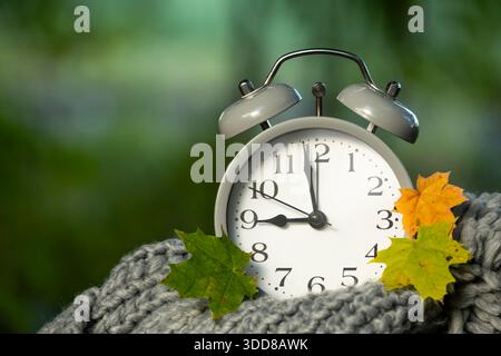 Eine Nahaufnahme eines klassischen Weckers, umgeben von bunten Herbstblättern, auf einem weichen, gestrickten Schal mit verschwommenem grünem Hintergrund. Stockfoto