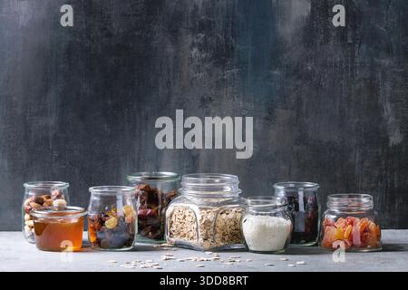 Auswahl an getrockneten Früchten, Nüssen, Honig und Haferflocken in verschiedenen Gläsern zum Kochen hausgemachter gesunder Frühstücksmüsli oder Müsli Energieriegel auf graublauem Tisch. Stockfoto