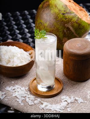 Ein hohes Glas Kokoswasser mit Eiswürfeln, Minzblättern und geschredderter Kokosnuss auf einem hölzernen Untersetzer, begleitet von einer Kokosnuss und einer Schüssel mit geschreddertem kokosnuss Stockfoto
