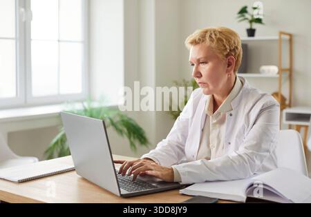 Senior fokussierte ernsthafte Ärztin in weißer Uniform, die online am Schreibtisch sitzt und am Computer arbeitet Stockfoto