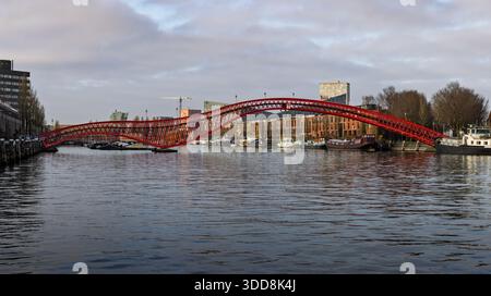 Die markante Python Bridge aus rotem Stahl überspannt das Wasser in den östlichen Docklands von Amsterdam. Moderne Fußgängerarchitektur verbindet Inseln. Stockfoto