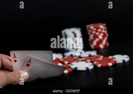 Hand hält ein Paar Asse, mit gestapelten und verstreuten Pokerchips auf dunklem Hintergrund Stockfoto