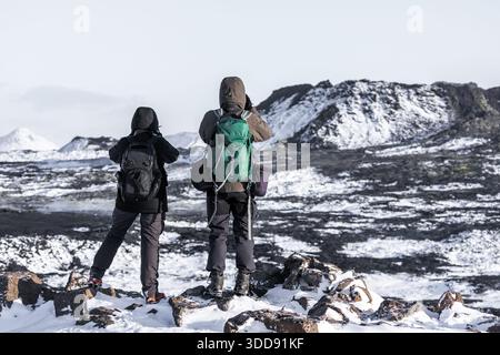 Grindavik, Reykjanes-Halbinsel, Island – 6. Februar 2022: Zwei Wanderer stehen auf schneebedeckten Felsen und beobachten ein zerklüftetes vulkanisches Lavafeld mit Stockfoto