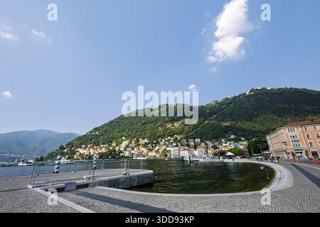 COMO, ITALIEN, 06.20.2025: Eine friedliche Promenade am See führt entlang eines ruhigen Hafens und zeigt bunte Hügelhäuser, grüne Berge und ein klares Blau Stockfoto