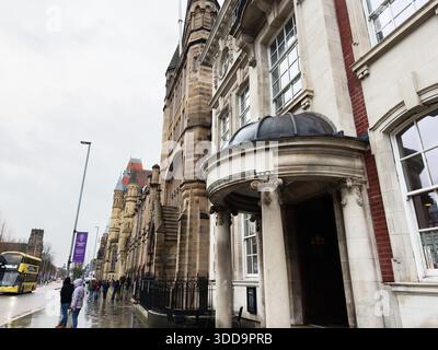 Manchester, Großbritannien - 06. Dezember 2025: Die Menschen gehen an einem großen Steingebäude in einer feuchten Manchester Street vorbei und beleuchten historische Architektur und Kultur Stockfoto