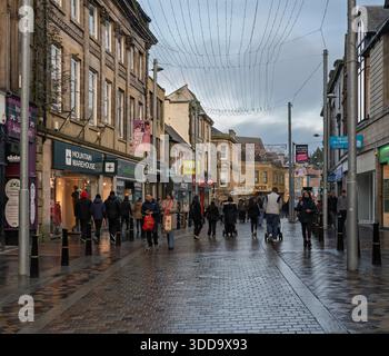 High Street in Inverness zeigt Menschen, die an einem bewölkten Dezembertag entlang der Straße laufen. Die Geschäfte sind geöffnet und es sind verschiedene Schilder zu sehen. Der Boden ist nass Stockfoto