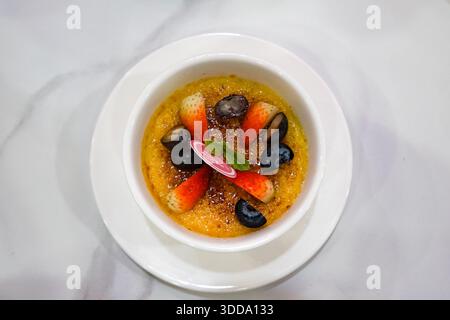 Elegante Creme Brulee in einem weißen Ramekin, garniert mit Erdbeeren, Heidelbeeren, Trauben, Minze und einer karamellisierten Zuckerkruste, serviert auf einer weißen Untertasse. Stockfoto