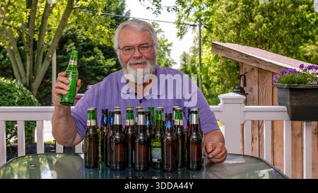 Ein Mann mit Bart sitzt draußen und hält eine grüne Flasche. Leere Bierflaschen stehen vor ihm auf einem Tisch und zeigen ein zwangloses Treffen in einem sonnigen Hinterhof. Stockfoto