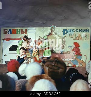 Slawjansk, Ukraine, UdSSR, 1980er Jahre Eine farbenfrohe Folklore-Vorstellung während des russischen Winterfestes auf der Bühne des nach Lenin benannten Parks (heute Shelkowitschy Park). Schauspieler in Kostümen von Ded Moroz, Narren und Tänzer treten für die Bürger auf. Die handgemalte Kulisse zeigt Illustrationen eines Samovars und Herds mit dem Text Russian Tea and Pancakes (Bliny). Bewohner in warmen Pelzhüten sehen die Show vom Publikum aus. Das Bild fängt die Freude an traditionellen Feiertagen und kulturellen Veranstaltungen in der friedlichen sowjetischen Stadt ein. Stockfoto