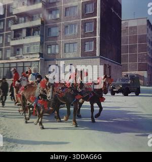 Slawjansk, Ukraine, UdSSR, 1980er Jahre Eine festliche Troika von Pferden zieht während der russischen Winterferien einen gemalten Holzschlitten durch das Artem-Mikroviertel. Schauspieler, die als Ded Moroz gekleidet sind, Snegurochka in einem Kokoshnik und helle Narren winken den Bewohnern zu. Der Folklorezug zieht vorbei an typischen sowjetischen Wohnhäusern und Fabrikschornsteinen. Ein Militärjeep der UAZ fährt in der Nähe. Die Szene steht in lebhaftem Kontrast zu traditionellen ländlichen Bräuchen und der modernen industriellen Stadtlandschaft der friedlichen Stadt. Stockfoto