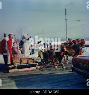 Slawjansk, Ukraine, UdSSR, 1980er Jahre Eine festliche Troika von Pferden zieht während der russischen Winterferien einen gemalten Holzschlitten durch das Artem-Mikroviertel. Schauspieler, die als Ded Moroz gekleidet sind, Snegurochka in einem Kokoshnik und helle Narren winken den Bewohnern zu. Der Folklorezug zieht vorbei an typischen sowjetischen Wohnhäusern und Fabrikschornsteinen. Ein Militärjeep der UAZ fährt in der Nähe. Die Szene steht in lebhaftem Kontrast zu traditionellen ländlichen Bräuchen und der modernen industriellen Stadtlandschaft der friedlichen Stadt. Stockfoto