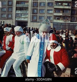 Slawjansk, Ukraine, UdSSR, 1980er Jahre Kostümierte Schauspieler posieren für ein Foto während des russischen Winterfestivals im Mikroviertel Artem. Ein lächelnder Snegurochka in einem traditionellen Kokoshnik steht neben einem Narren mit einem bemalten Schnurrbart und anderen Darstellern in weißen und roten Clown-Outfits. Im Hintergrund versammelt sich eine Menschenmenge von Einwohnern in der Nähe eines großen Wohngebäudes mit grauen Paneelen, um die Show zu sehen. Dieses Archivbild fängt die Atmosphäre des Amateurtheaters und die fröhlichen Gemeindefeiern im Wohnviertel der sowjetischen Stadt ein. Stockfoto