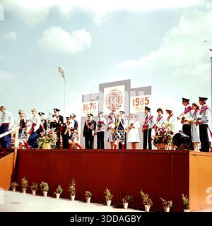 Slawjansk, Ukraine, UdSSR, 8. September 1985. Eine offizielle Zeremonie zum Stadttag findet auf einer Bühne am Oktyabrskoy Revolyutsii-Platz (heute Soborna-Platz) statt. Eine große Kulisse zeigt das Gründungsjahr 1676 und das laufende Jahr 1985. Veteranen des Zweiten Weltkriegs mit Medaillen und junge Ehrenbürger mit Schärpen stehen auf dem Podium, während ein Offizier am Mikrofon spricht. Tänzer in ukrainischen Trachten nehmen an der Veranstaltung teil. Dieses Archivbild fängt die patriotische Feier und Anerkennung der Bewohner der friedlichen Sowjetstadt ein. Stockfoto