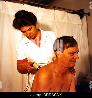 Slawjansk, Ukraine, UdSSR, 1980er Jahre Eine Krankenschwester führt ein physiotherapeutisches Verfahren an einem männlichen Patienten im Bogatyr-Präventivorium des Khimprom-Werks durch. Der Mediziner in weißer Uniform verwendet ein Handgerät, um den Rücken und die Schultern des Mannes zu behandeln. Die Szene findet in einem Bestrahlungsraum mit weißen Vorhängen statt. Diese Einrichtung bot kostenlose Gesundheitsversorgung, Massagen und Rehabilitation für industrielle Mitarbeiter. Das Archivfoto zeigt die Qualität der medizinischen Versorgung und Versorgung der Arbeiterklasse in der friedlichen Sowjetstadt. Stockfoto