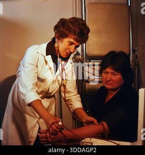 Slawjansk, Ukraine, UdSSR, 1980er Jahre Eine Ärztin misst den Blutdruck eines Patienten im Bogatyr-Präventivorium der Khimprom-Anlage. Der Arzt in einem weißen Fell verwendet ein manuelles Sphygmomanometer und Stethoskop, um die Gesundheit der Frau während einer Routineuntersuchung zu überprüfen. Die Szene findet in einem mit Rehabilitationsgeräten ausgestatteten medizinischen Raum statt. An diesem Standort wurden regelmäßige Kontrollen und Behandlungen für Industriearbeiter durchgeführt. Das Archivbild zeigt die aufmerksame medizinische und soziale Unterstützung der Mitarbeiter in der friedlichen Sowjetstadt. Stockfoto