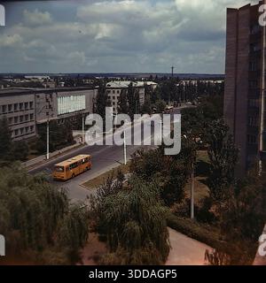 Slawjansk, Ukraine, UdSSR, 1980er Jahre Panoramablick auf die Yunykh Kommunarov Straße (heute Bankowskaja Straße) im Stadtteil SATU. Das große modernistische Gebäude der Technischen Hochschule für Zivilluftfahrt steht auf der linken Seite und ist mit einem sowjetischen Wandgemälde geschmückt. Klassische öffentliche Verkehrsmittel, darunter ein gelber LIAZ und ein rot-weißer Ikarus-Bus, fahren entlang der breiten Allee, die von grünen Pappeln und Weiden gesäumt ist. In der Ferne eröffnet sich der Blick auf die Lozanovich-Straße und den Khytry-Markt. Dieses Archivbild erfasst die Stadtplanung, den Verkehr und den Rhythmus des friedlichen Donbas Stockfoto