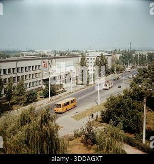 Slawjansk, Ukraine, UdSSR, 1980er Jahre Panoramablick auf die Yunykh Kommunarov Straße (heute Bankowskaja Straße) im Stadtteil SATU. Das große modernistische Gebäude der Technischen Hochschule für Zivilluftfahrt steht auf der linken Seite und ist mit einem sowjetischen Wandgemälde geschmückt. Klassische öffentliche Verkehrsmittel, darunter ein gelber LIAZ und ein rot-weißer Ikarus-Bus, fahren entlang der breiten Allee, die von grünen Pappeln und Weiden gesäumt ist. In der Ferne eröffnet sich der Blick auf die Lozanovich-Straße und den Khytry-Markt. Dieses Archivbild erfasst die Stadtplanung, den Verkehr und den Rhythmus des friedlichen Donbas Stockfoto