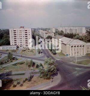 Slawjansk, Ukraine, UdSSR, 1980er Jahre Ein Panoramablick aus einem hohen Winkel mit Blick auf die landwirtschaftliche Technische Hochschule in der Chubar-Straße (heute Swetlodarskaya-Straße). Das Bildungsgebäude verfügt über einen klassischen Säulengang, der im Kontrast zum modernen neunstöckigen Wohnblock aus weißem Backstein auf der anderen Straßenseite steht. Die breiten Straßen sind ruhig mit minimalem Verkehr, umgeben von grünen Rasenflächen und jungen Tannen. Dieses Archivbild erfasst die Stadtplanung, die Bildungsinfrastruktur und die friedliche Atmosphäre der sowjetischen Donbass-Stadt. Stockfoto