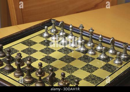 Ein feines Schachspiel auf einer gelben Pinnwand zieht die Aufmerksamkeit auf sich. Metallische Figuren in Silber und Bronze befinden sich in einer perfekten Ausgangsposition und reflektieren Stockfoto