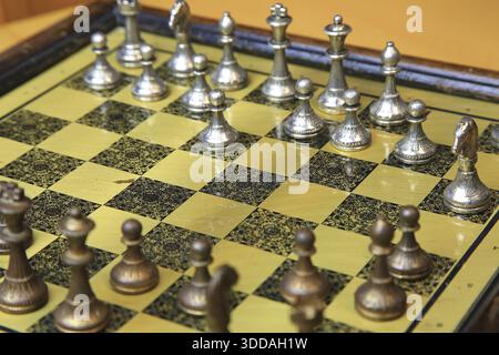 Ein feines Schachspiel auf einer gelben Pinnwand zieht die Aufmerksamkeit auf sich. Metallische Figuren in Silber und Bronze befinden sich in einer perfekten Ausgangsposition und reflektieren Stockfoto