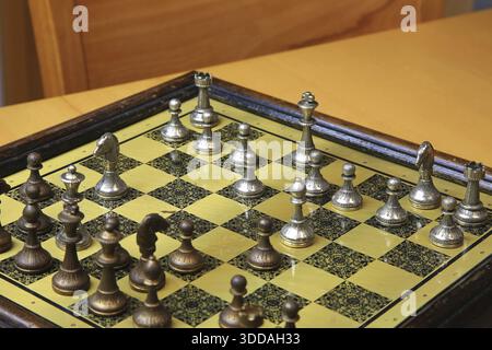 Ein feines Schachspiel auf einer gelben Pinnwand zieht die Aufmerksamkeit auf sich. Metallische Figuren in Silber und Bronze befinden sich in einer perfekten Ausgangsposition und reflektieren Stockfoto