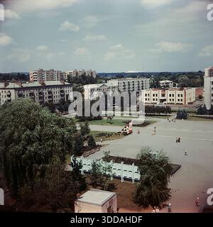 Slawjansk, Ukraine, UdSSR, 1980er Jahre Erhöhter Panoramablick auf das Stadtzentrum vom Steuergebäude aus. Die Aufnahme erfasst den Platz der Oktoberrevolution (heute Soborna Square) und die Lenin Street (heute University St). Sowjetische Propaganda-Slogans "Frieden für die Welt" (Miru mir) und "Ruhm für die KPdSU" (Slava KPSS) sind auf den Dächern von Wohnziegelbauten zu sehen. In der Gegend gibt es üppiges Grün, große Weiden und Bürger, die entlang der Alleen spazieren. (558 знаков — идеально, почти предел). Stockfoto