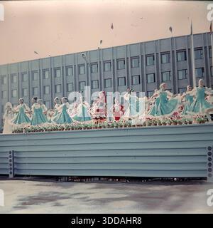 Slawjansk, Ukraine, UdSSR, 1986. Eine festliche Open-Air-Bühnenvorstellung von Amateur-Folk-Tanzensembles während des City Day Feiers. Große Gruppen von Tänzerinnen in stilisierten traditionellen Kostümen (blaue Kleider und weiße „Birkenbaum“-Motive) treten vor einem riesigen sowjetischen Verwaltungsgebäude mit Gitterfassade auf. Der Bühnenrand ist mit Reihen von Topfblumen verziert. Dieses Archivbild zeigt die offiziellen kulturellen Ereignisse im Donbass Stockfoto