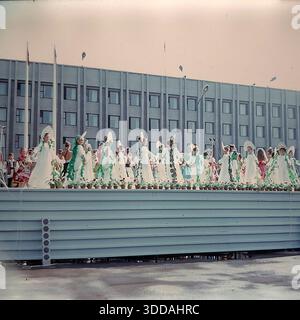 Slawjansk, Ukraine, UdSSR, 1986. Eine festliche Open-Air-Bühnenvorstellung von Amateur-Folk-Tanzensembles während des City Day Feiers. Große Gruppen von Tänzerinnen in stilisierten traditionellen Kostümen (blaue Kleider und weiße „Birkenbaum“-Motive) treten vor einem riesigen sowjetischen Verwaltungsgebäude mit Gitterfassade auf. Der Bühnenrand ist mit Reihen von Topfblumen verziert. Dieses Archivbild zeigt die offiziellen kulturellen Ereignisse im Donbass Stockfoto
