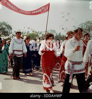 Slawjansk, Ukraine, UdSSR, 1980er Jahre Eine festliche Prozession eines Amateurchors oder Volksensembles während eines Kulturfestivals auf dem Platz der Oktoberrevolution. Die Gruppe besteht aus Männern und Frauen in stilisierten rot-weißen Bühnenkostümen, die mit großen Blumenmustern verziert sind. Sie tragen Äste mit künstlichen weißen Blüten, die den Frühling symbolisieren. Ein rotes Banner mit dem pazifistischen Slogan „...NUZHEN ... PLANETE!“ (Der Planet braucht Frieden) schwimmt über der Säule. Dieses Archivbild fängt das offizielle kulturelle Leben und die Friedenspropaganda der Sowjetzeit ein Stockfoto