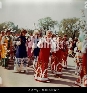 Slawjansk, Ukraine, UdSSR, 1980er Jahre Eine farbenfrohe Prozession von Amateur-Volkskunst-Kollektiven während eines Kulturfestivals auf dem Platz der Oktoberrevolution. Frauen eines Volkschors tragen lange rote, blaue und gelbe Bühnenkleider mit großen Blumenornamenten. Sie tragen Äste mit künstlichen weißen Blüten und Ballons, die den Frühling symbolisieren. Dieses Archivbild zeigt die kulturellen Massenveranstaltungen, das Kostümdesign und die festliche Atmosphäre der sowjetischen Ära im Donbass. Stockfoto