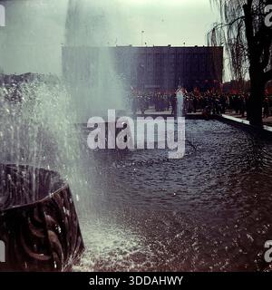 Slawjansk, Ukraine, UdSSR, 1980er Jahre Blick auf den funktionierenden Brunnen auf dem zentralen Platz der Oktoberrevolution (heute Soborna-Platz) während einer Großstadtfeier. Im Vordergrund sprühen Wasserstrahlen aus einer Struktur mit dekorativen Metallgittern nach oben. Im Hintergrund versammelt sich eine große Menschenmenge mit roten Fahnen vor dem sowjetischen Verwaltungsgebäude (Stadtrat). Dieses Archivbild fängt die festliche Atmosphäre und die urbane Landschaft der Donbass-Stadt ein. Stockfoto