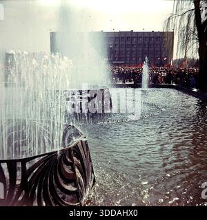 Slawjansk, Ukraine, UdSSR, 1980er Jahre Blick auf den funktionierenden Brunnen auf dem zentralen Platz der Oktoberrevolution (heute Soborna-Platz) während einer Großstadtfeier. Im Vordergrund sprühen Wasserstrahlen aus einer Struktur mit dekorativen Metallgittern nach oben. Im Hintergrund versammelt sich eine große Menschenmenge mit roten Fahnen vor dem sowjetischen Verwaltungsgebäude (Stadtrat). Dieses Archivbild fängt die festliche Atmosphäre und die urbane Landschaft der Donbass-Stadt ein. Stockfoto