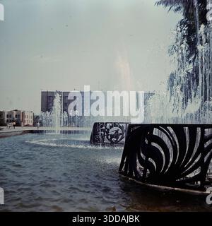 Slawjansk, Ukraine, UdSSR, 1980er Jahre Blick auf den funktionierenden Brunnen auf dem zentralen Platz der Oktoberrevolution (heute Soborna-Platz) während einer Großstadtfeier. Im Vordergrund sprühen Wasserstrahlen aus einer Struktur mit dekorativen Metallgittern nach oben. Im Hintergrund versammelt sich eine große Menschenmenge mit roten Fahnen vor dem sowjetischen Verwaltungsgebäude (Stadtrat). Dieses Archivbild fängt die festliche Atmosphäre und die urbane Landschaft der Donbass-Stadt ein. Stockfoto
